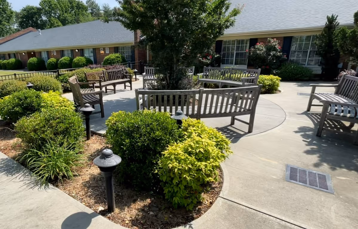 Outdoor seating area with multiple wooden benches arranged around a circular concrete pathway, surrounded by green bushes and plants. In the background, there is a single-story brick building with windows and a gray roof under a clear sky.