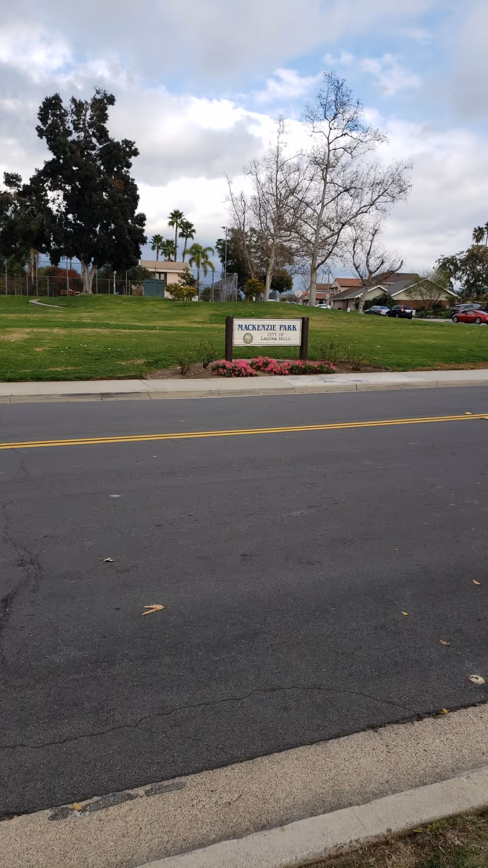 A grassy park area with a sign that reads 'Mackenzie Park City of Laguna Hills' surrounded by flowers. There are trees, houses, and parked cars in the background under a partly cloudy sky.