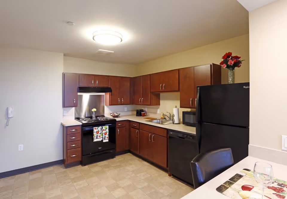A modern kitchen with dark wood cabinets, a black refrigerator, black dishwasher, black stove with a stainless steel backsplash, and a beige countertop. There is a vase with red and pink flowers on top of the refrigerator, a paper towel holder, a microwave, and a small cutting board near the sink. A dining area with a chair and placemat is partially visible in the foreground.