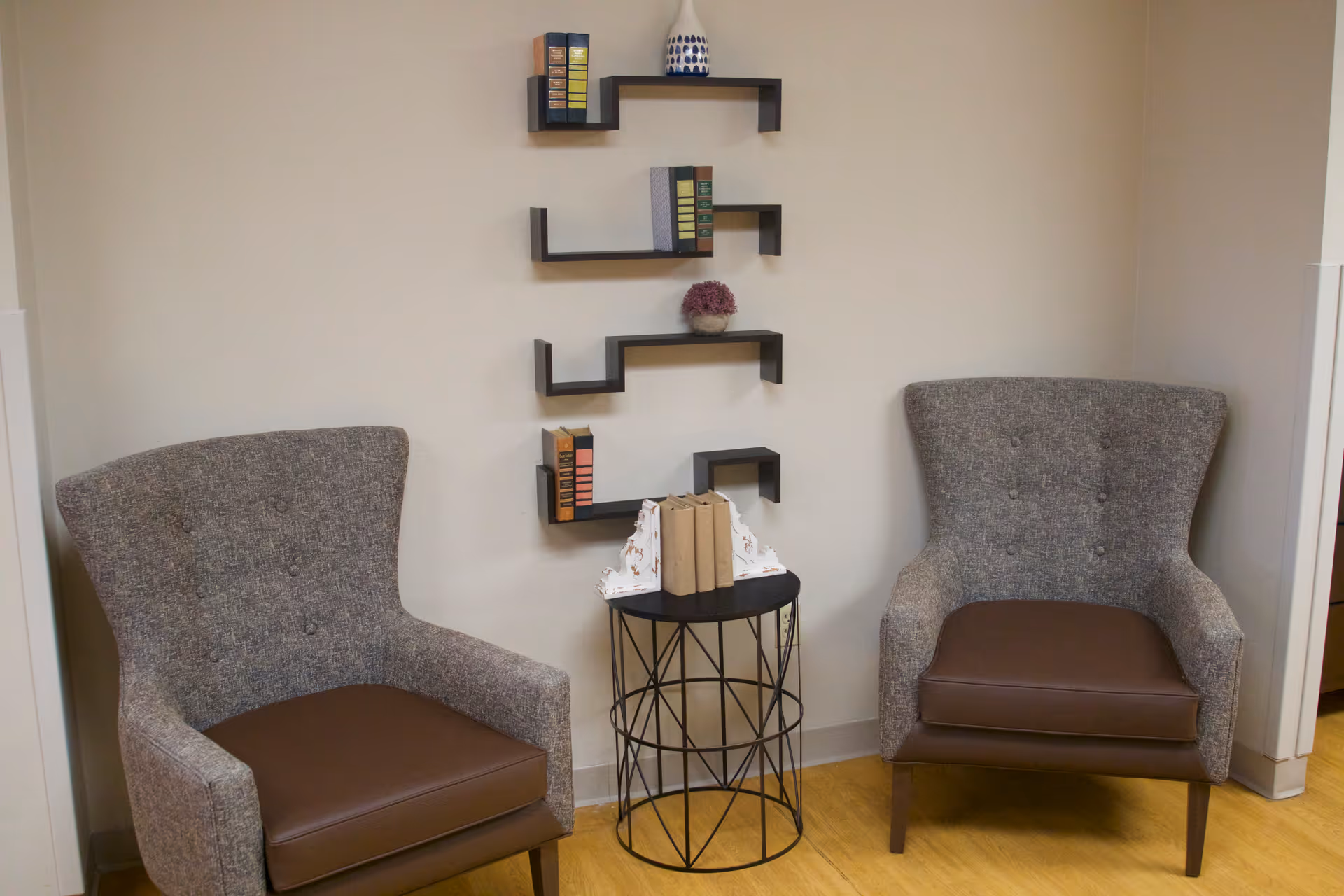 Two upholstered armchairs flanking a small round side table beneath wall-mounted zigzag shelves holding books and decor.