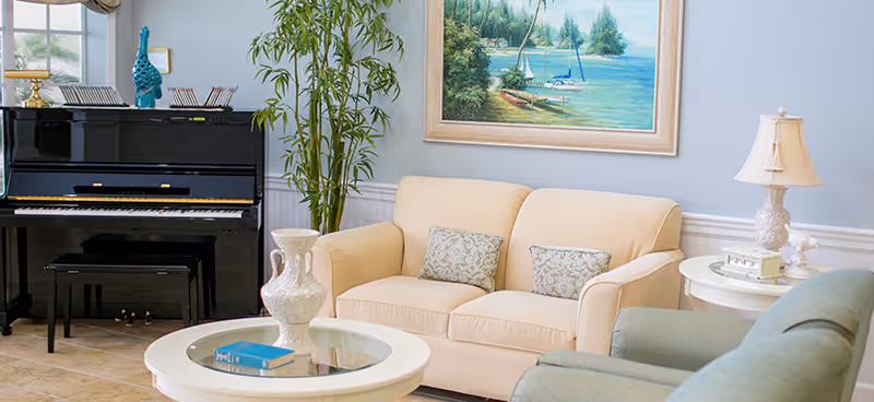 Cozy living room with a beige loveseat, armchairs, round glass-topped coffee table, upright piano, and a coastal painting on the wall.