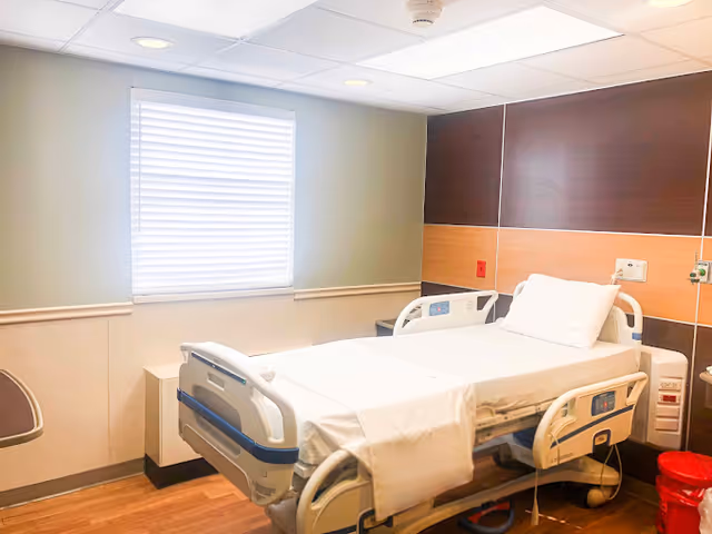 A clean hospital patient room with an adjustable bed, pillow, window with blinds, and medical equipment.