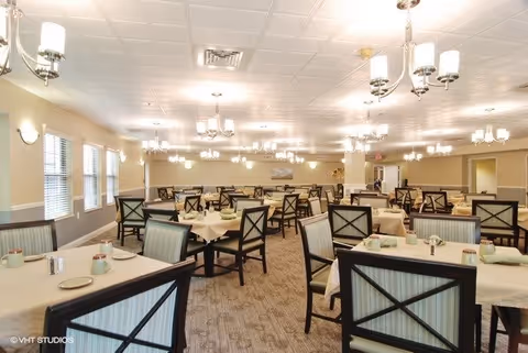 A spacious dining room with multiple tables covered in beige tablecloths, each set with plates, cups, and silverware. The room features cushioned chairs with wooden frames, large windows on one side, and multiple ceiling light fixtures providing bright illumination.