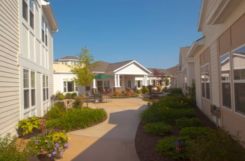 Outdoor courtyard area of Glenmere Assisted Living with a paved walkway, landscaped garden beds, and seating areas under umbrellas, surrounded by light-colored buildings under a clear blue sky.