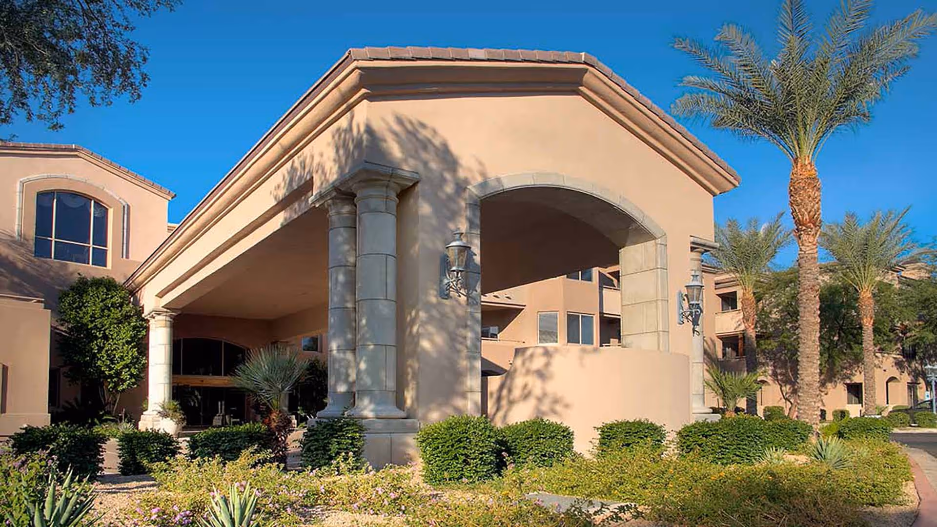 Entrance of a beige stucco senior living building with large pillars, palm trees, and landscaped shrubs under a clear blue sky.