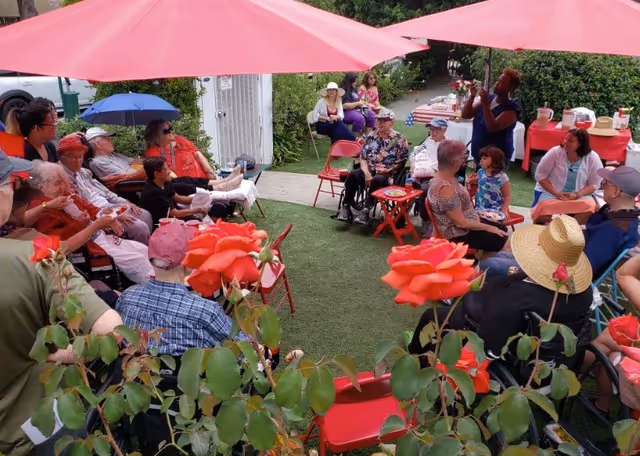 A group of elderly people and caregivers gathered outdoors under large pink umbrellas, sitting on red chairs arranged in a circle on a lawn. Some people are in wheelchairs, and there are red roses in the foreground. A woman is standing and speaking to the group, while others listen attentively. There is a table with refreshments in the background.