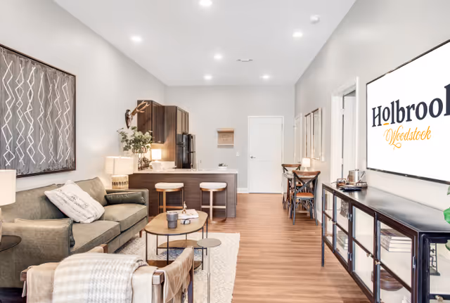 Open-plan living area with a sofa and armchair, coffee table, kitchen island with two stools, and a wall sign reading 'Holbrook Woodstock'.