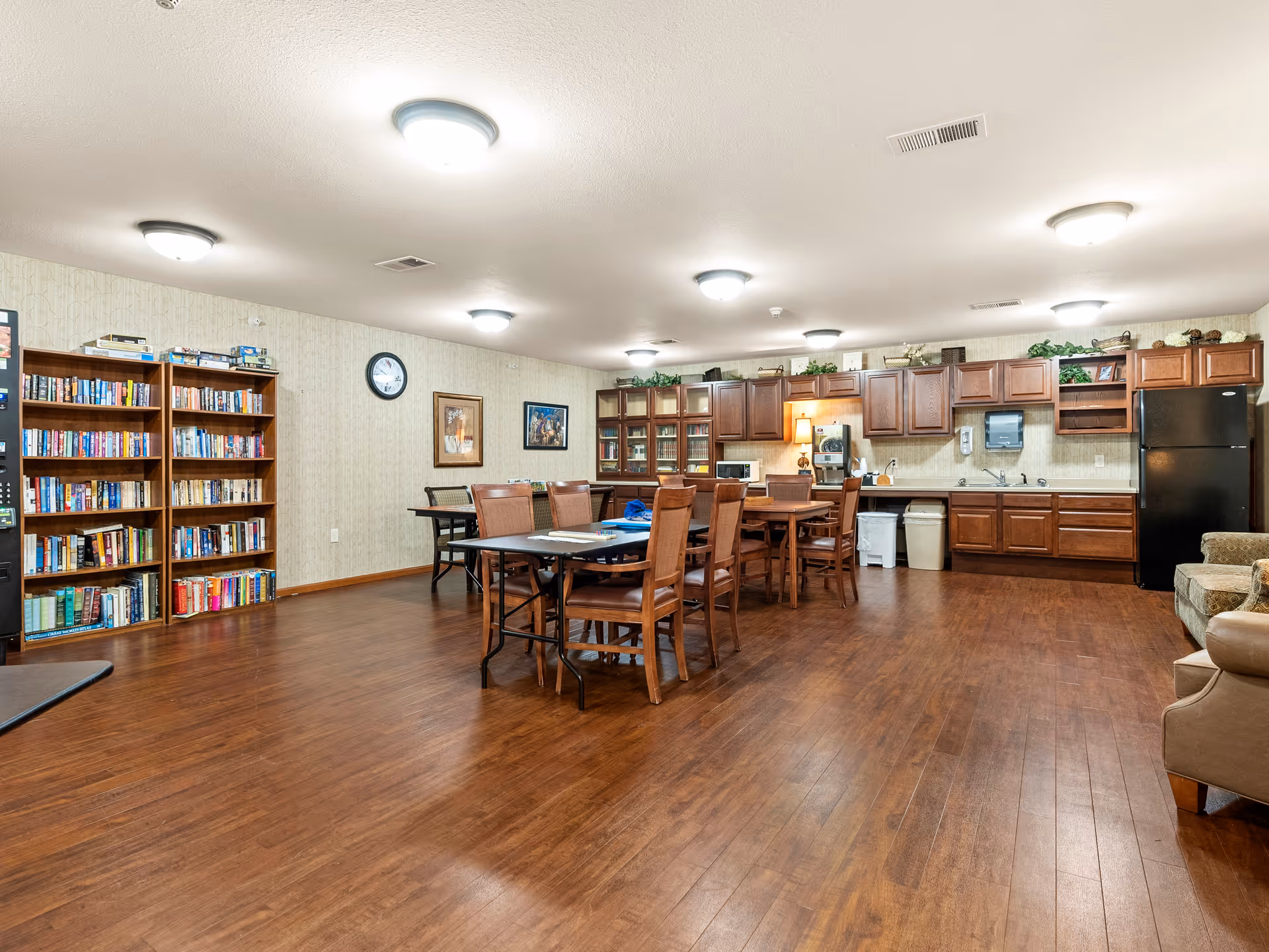 Communal senior living common room with dining tables and chairs, bookshelves, and a kitchenette with cabinets and refrigerator.