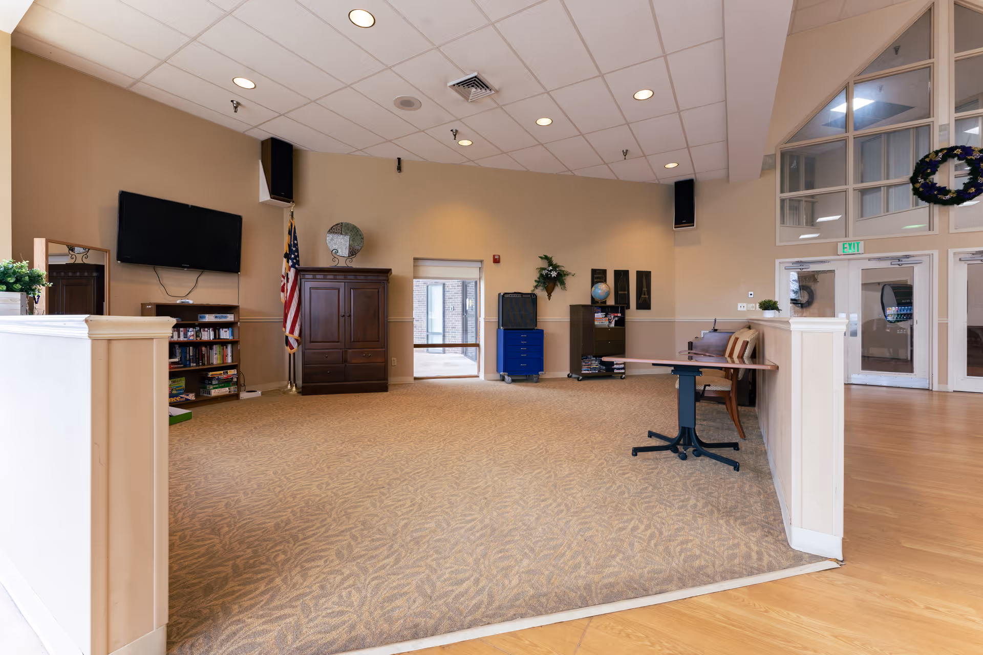 A spacious common area in a senior living facility with beige walls and carpeted floor. The room features a wall-mounted flat screen TV, an American flag, a wooden cabinet, bookshelves with books and games, a small table with chairs, and large windows and doors leading to another area. The ceiling has recessed lighting and speakers.