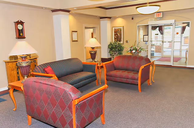 A seating area in a senior living facility lobby featuring three upholstered chairs and a sofa with wooden frames, two table lamps on side tables, a wall clock, framed artwork, potted plants, and glass entrance doors in the background.