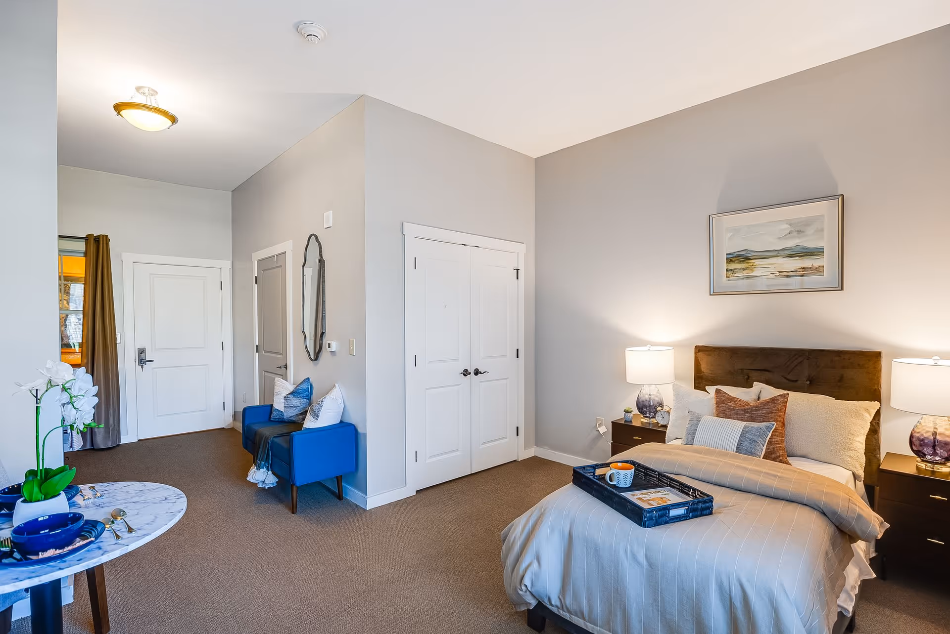 A cozy bedroom in a senior living facility with a neatly made bed featuring multiple pillows and a tray with a cup and book. Two bedside tables with matching lamps flank the bed. A framed landscape painting hangs above the headboard. To the left, there is a small sitting area with two blue chairs and decorative pillows, a wall mirror, and a white double-door closet. A round table with a blue bowl and white orchid is in the foreground. The room has beige carpeting and light gray walls.
