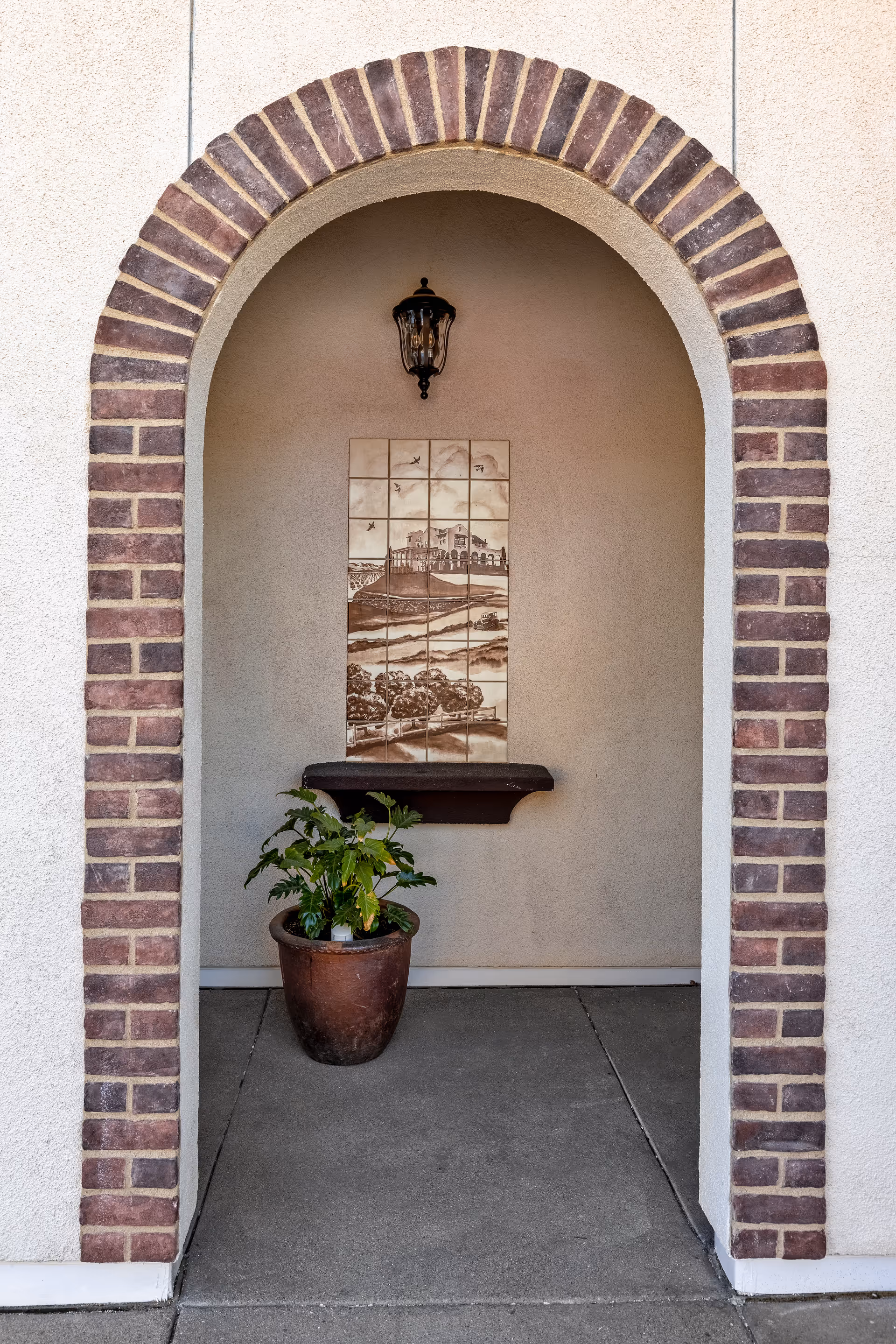 Arched brick entry alcove with a wall lantern, decorative tile mural, small shelf and a potted plant.