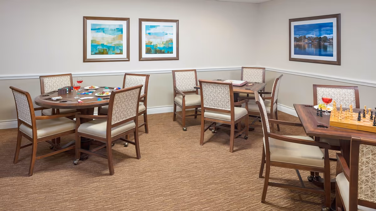 A cozy room with three wooden tables surrounded by cushioned chairs. Two tables have plates with snacks and drinks, while the third table has a chessboard set up for a game. The walls are decorated with three framed pictures, and the room has beige carpeting and light-colored walls with white trim.