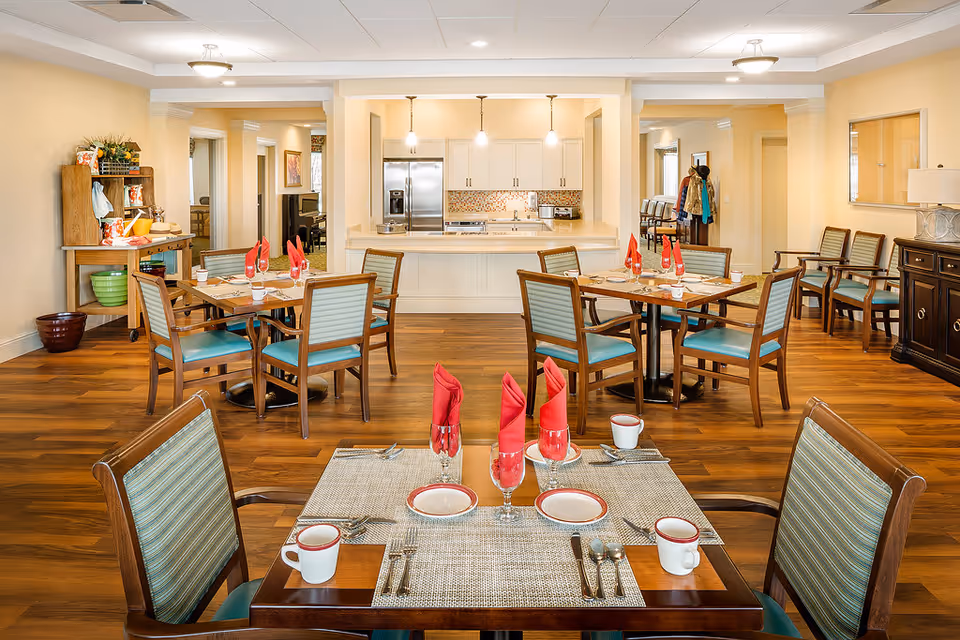 A bright dining room in a retirement community with wooden floors and several tables set for meals. Each table has place settings with plates, cups, utensils, and red folded napkins. The room features a kitchen area in the background with white cabinets, a refrigerator, and pendant lights. There are chairs with green cushions around the tables and additional seating along the walls. The space is warmly lit and decorated with a few plants and artwork.