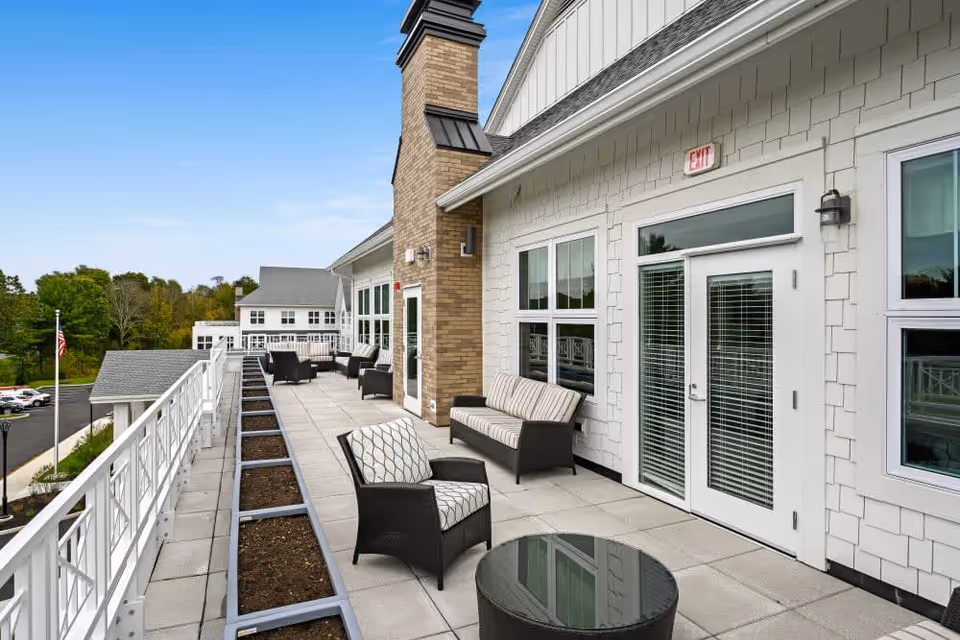 Outdoor balcony area at Riverbend at Farmington with cushioned wicker chairs and sofas arranged along the wall. The balcony has a railing on one side and planter boxes along the edge. The building exterior features white siding and large windows, with a brick chimney in the center. The sky is clear and blue, and there is a parking lot and trees visible in the background.