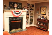 Cozy living room area with a white fireplace decorated with a red, white, and blue bunting. Above the fireplace is a framed circular artwork, and the mantel holds several decorative items. To the right of the fireplace is a built-in white shelving unit filled with framed photos and decorative objects. A small wooden cart with framed pictures and a lamp is positioned against the wall. The room has warm lighting and a carpeted floor.