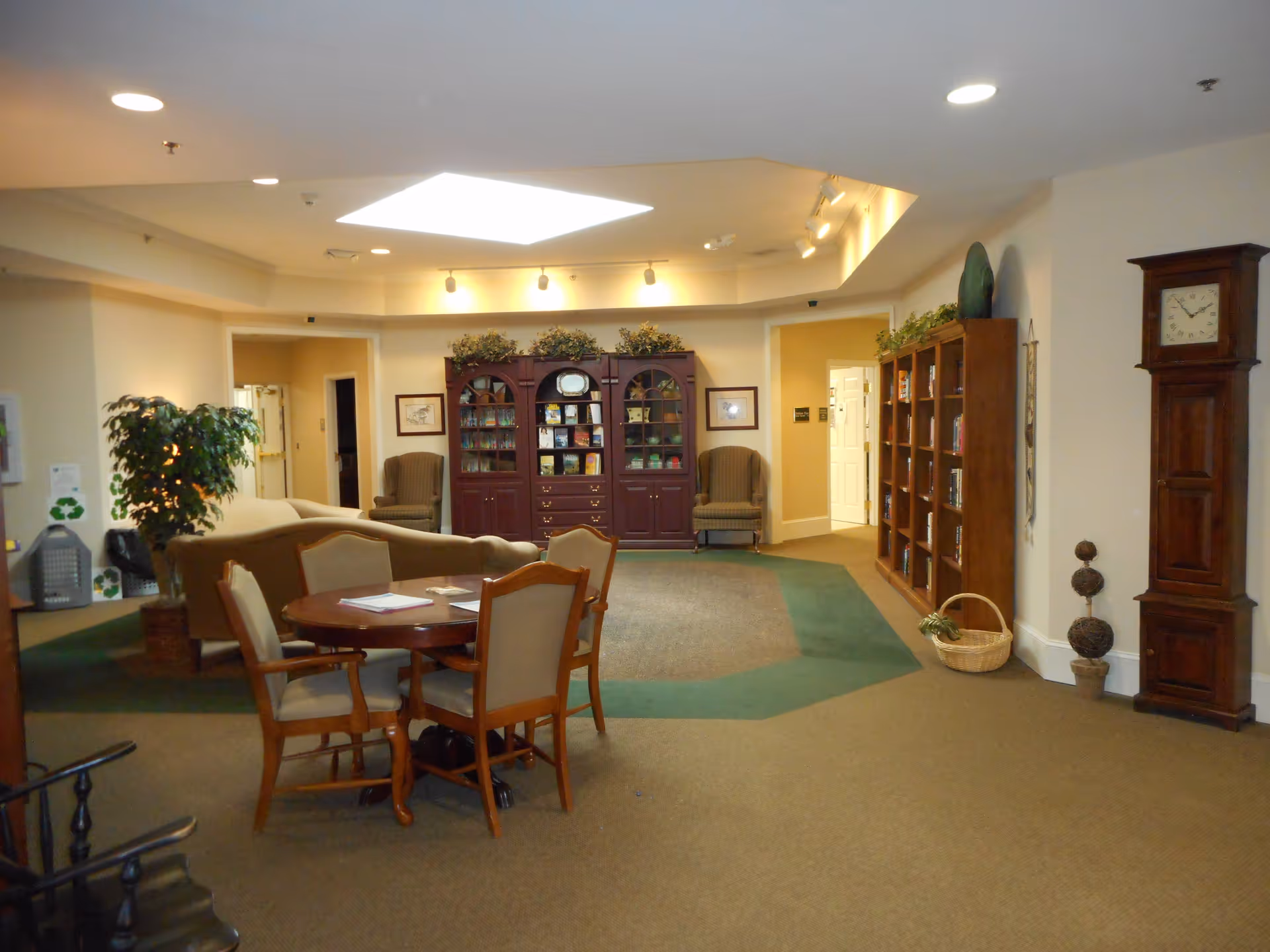 A cozy common area in a senior living facility featuring a round wooden table with four upholstered chairs, a beige sofa, two armchairs, and multiple bookshelves filled with books and decorative items. The room has a large skylight, recessed lighting, a tall wooden grandfather clock, and plants for decoration.