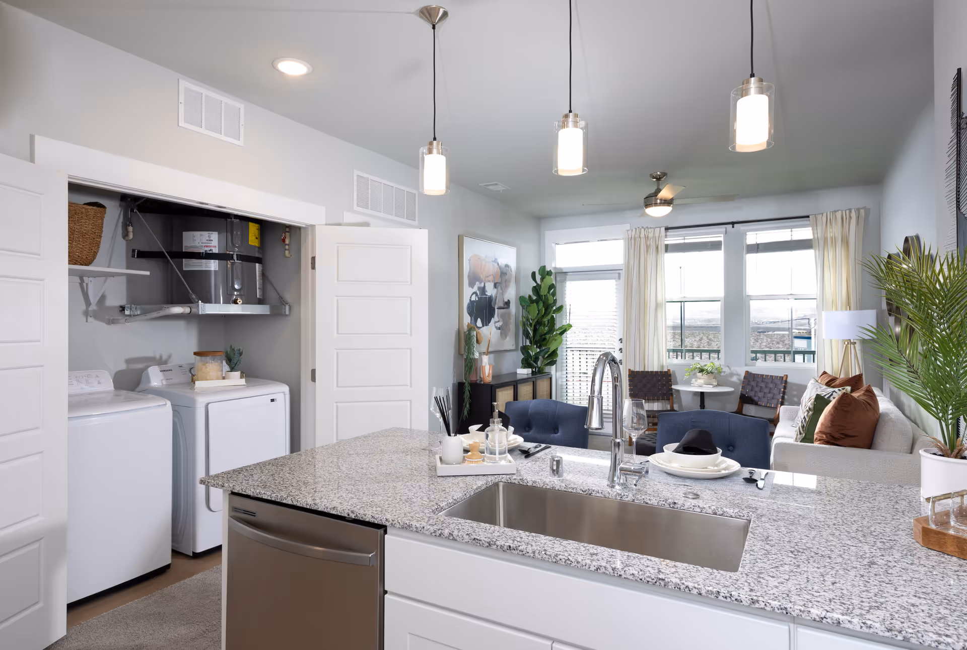 Modern open kitchen with a granite countertop island featuring a sink and faucet, set with plates and utensils. Behind the kitchen is a laundry area with a washer and dryer, and a water heater above them. The living room area is visible in the background with a sofa, chairs, plants, and large windows with curtains letting in natural light.