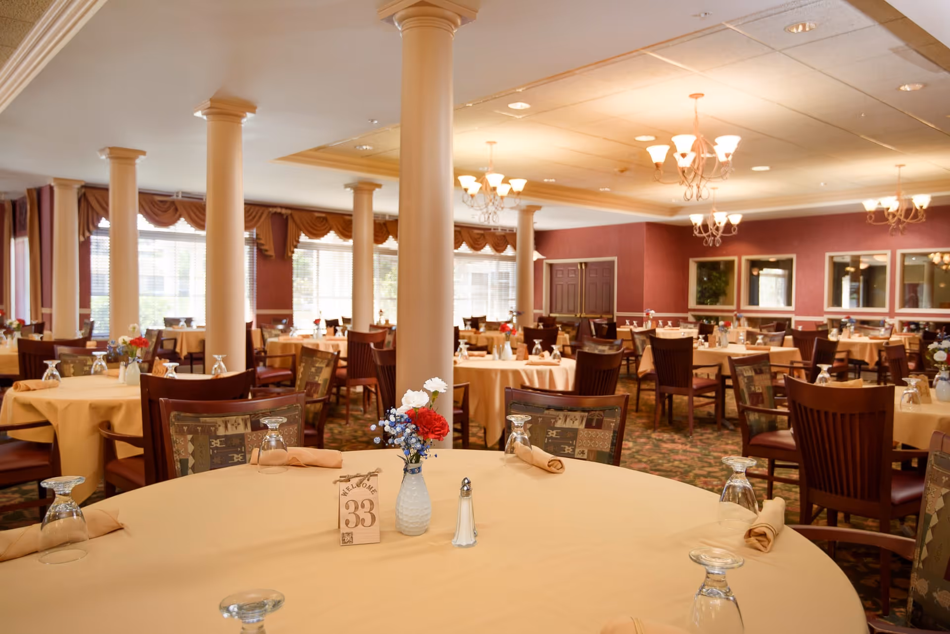 A spacious dining room with round tables covered in beige tablecloths, each set with upside-down glasses, rolled napkins, and small flower vases. The room features multiple white columns, large windows with brown valances, chandeliers, and a patterned carpet.