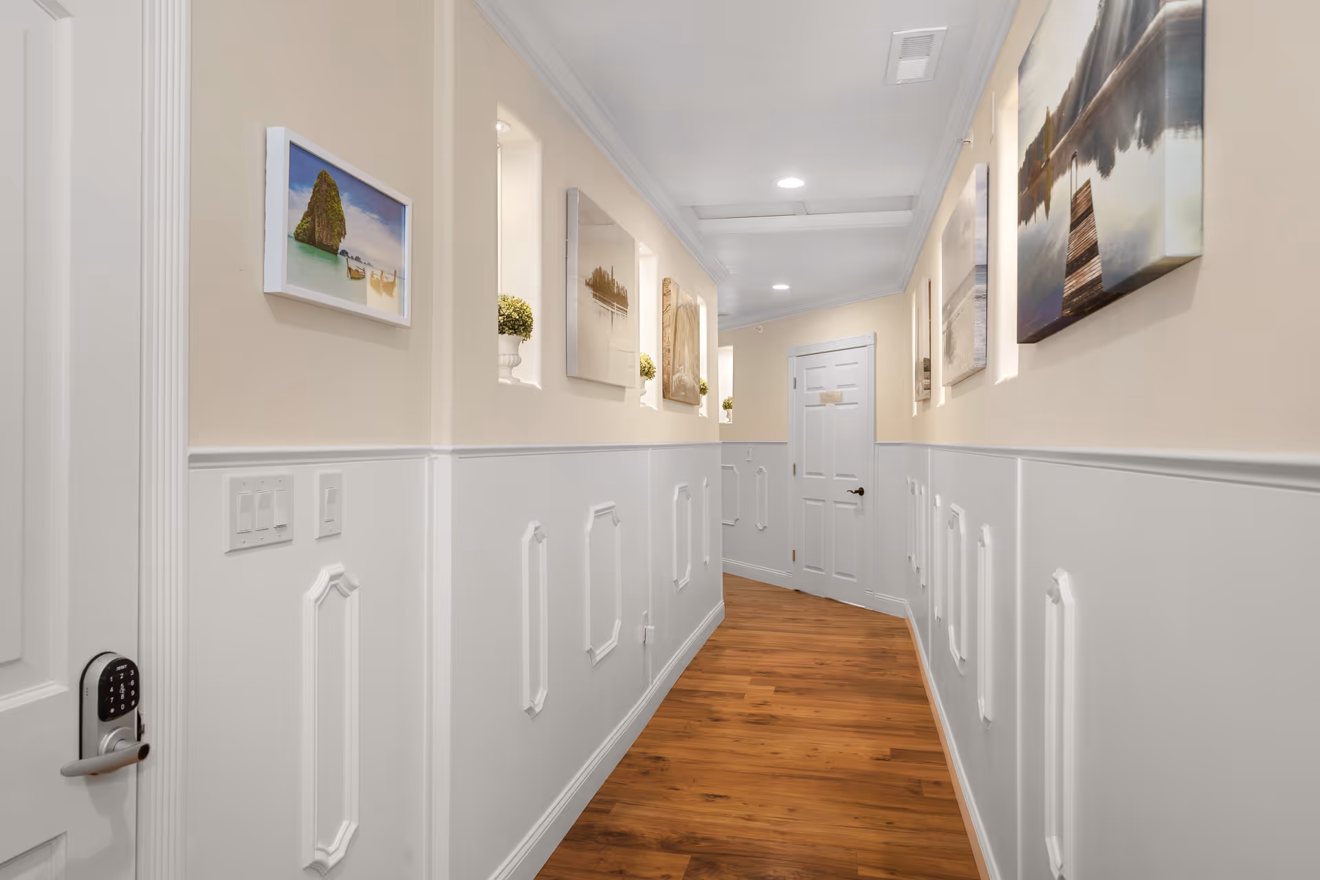 A well-lit hallway with wooden flooring and white wainscoting on the walls. The upper walls are painted beige and decorated with framed landscape artwork and small potted plants in recessed wall niches. There is a white door at the end of the hallway and a keypad lock on the door to the left.