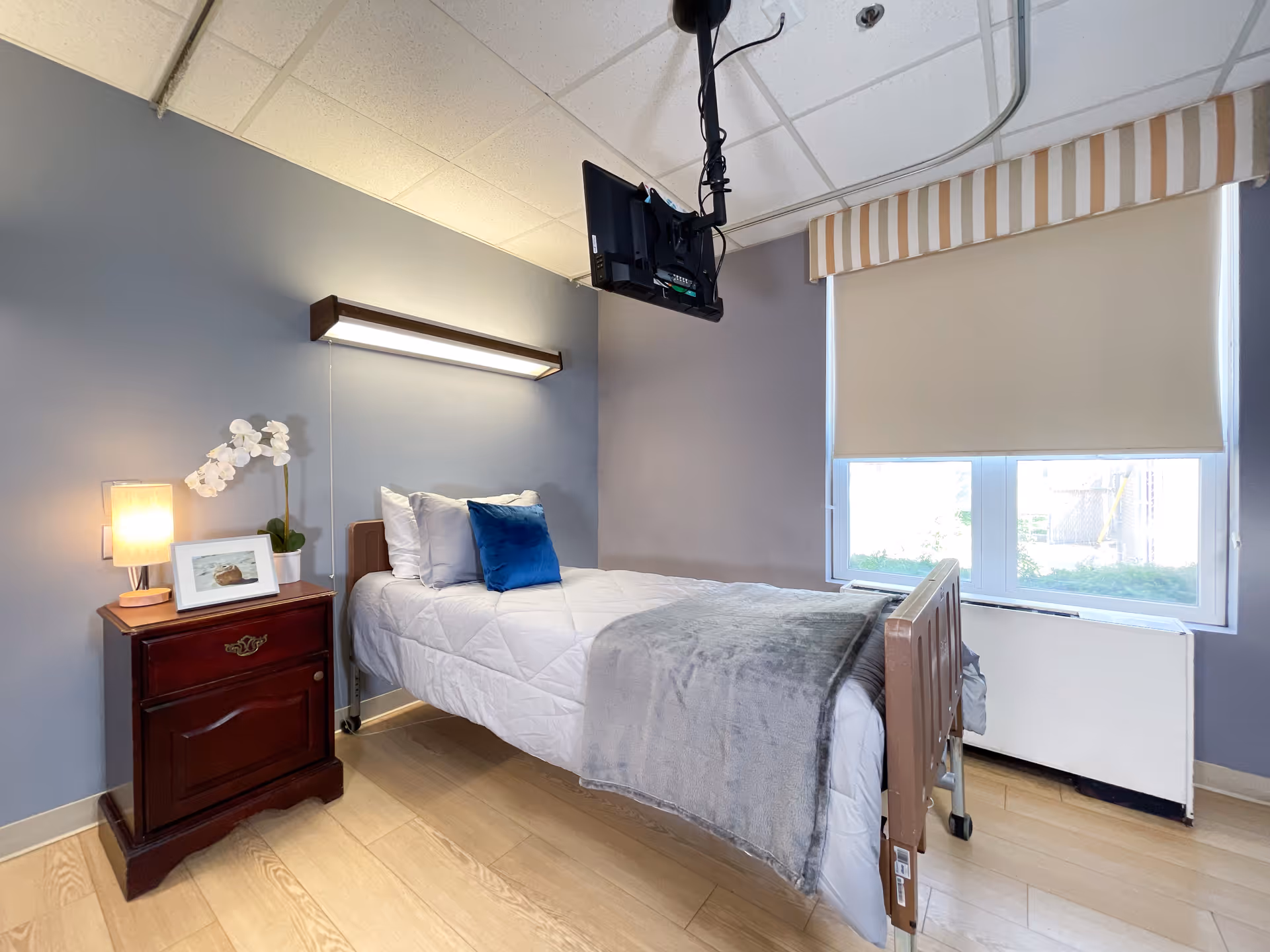 A tidy senior care bedroom with a single hospital-style bed, bedside table and lamp, ceiling-mounted TV, and a window with a blind.
