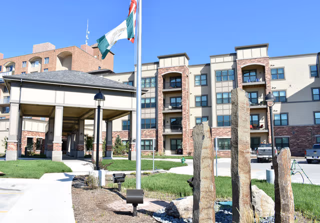 Exterior view of a multi-story senior living facility with a covered entrance, flagpoles with flags, landscaped grounds, and stone sculptures in the foreground under a clear blue sky.