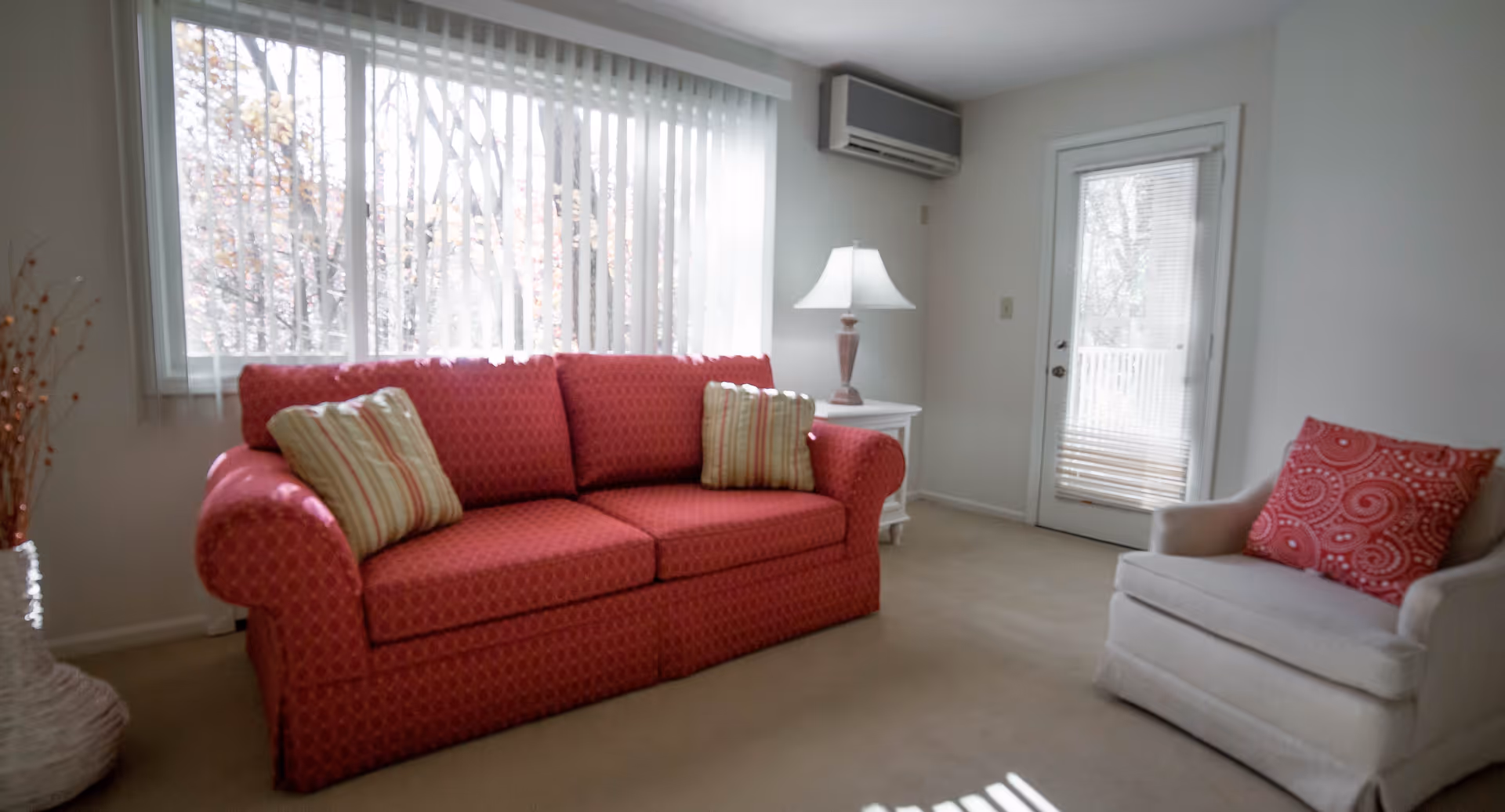 A cozy living room with a red patterned sofa adorned with striped cushions, a beige armchair with a red patterned pillow, a white side table with a lamp, vertical blinds covering a large window, and a glass door leading outside.