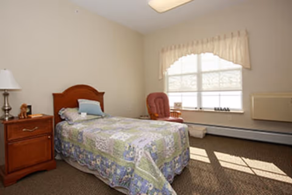 A simple bedroom with a single bed covered in a patchwork quilt, a wooden nightstand with a lamp and small decorative items, a red cushioned chair near a large window with blinds and a valance, and beige walls and carpeted floor.