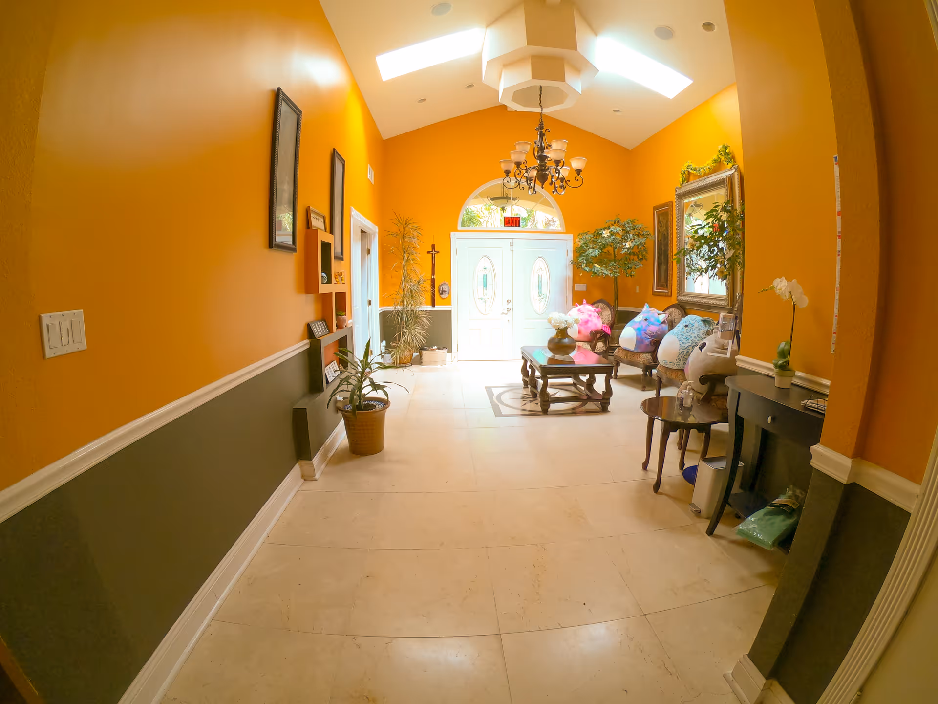 A bright hallway with orange and dark green walls, cream-colored tiled floor, and a high ceiling with skylights and a chandelier. The hallway features a wooden coffee table with a flower vase, several chairs with colorful cushions, potted plants, framed pictures, and a large mirror. Double doors with oval glass panels are visible at the end of the hallway.