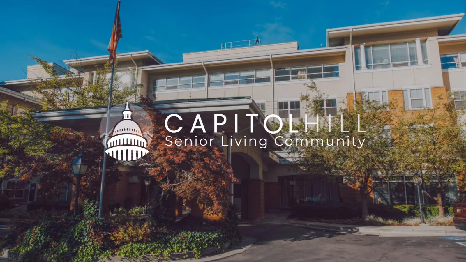 Exterior front view of Capitol Hill Senior Living Community building with trees, a flagpole, and a driveway under a clear blue sky.