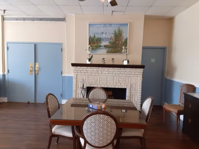 A cozy interior room featuring a square wooden table with a glass top surrounded by four patterned chairs. Behind the table is a white brick fireplace with a wooden mantle, decorated with small plants and a landscape painting above it. The room has light-colored walls with blue double doors on the left and a single blue door on the right. There is also a wooden chair and a dark wooden cabinet against the right wall. The floor is wooden, and the ceiling has white tiles with a ceiling fan.