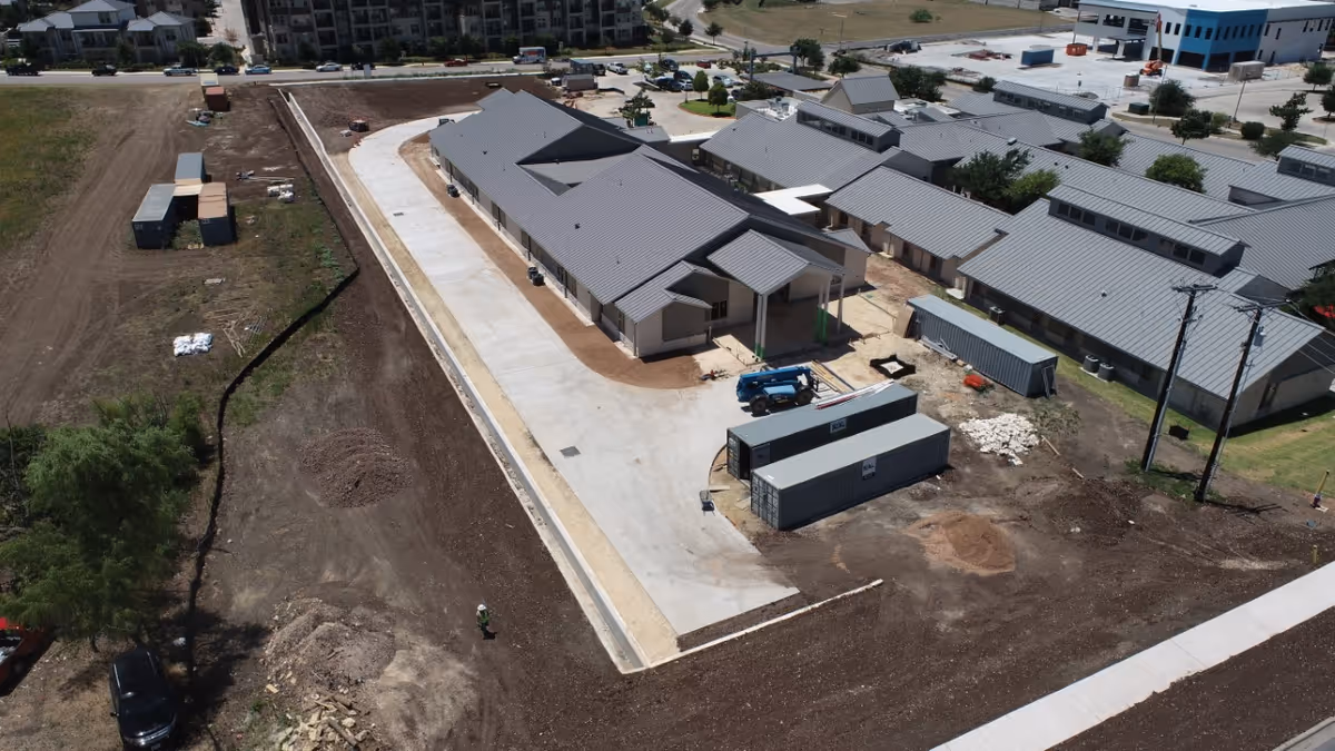 Aerial view of a construction site at University Village showing multiple single-story buildings with gray metal roofs, surrounded by dirt and construction materials. There are several shipping containers and construction equipment on site, with a paved driveway running through the area.