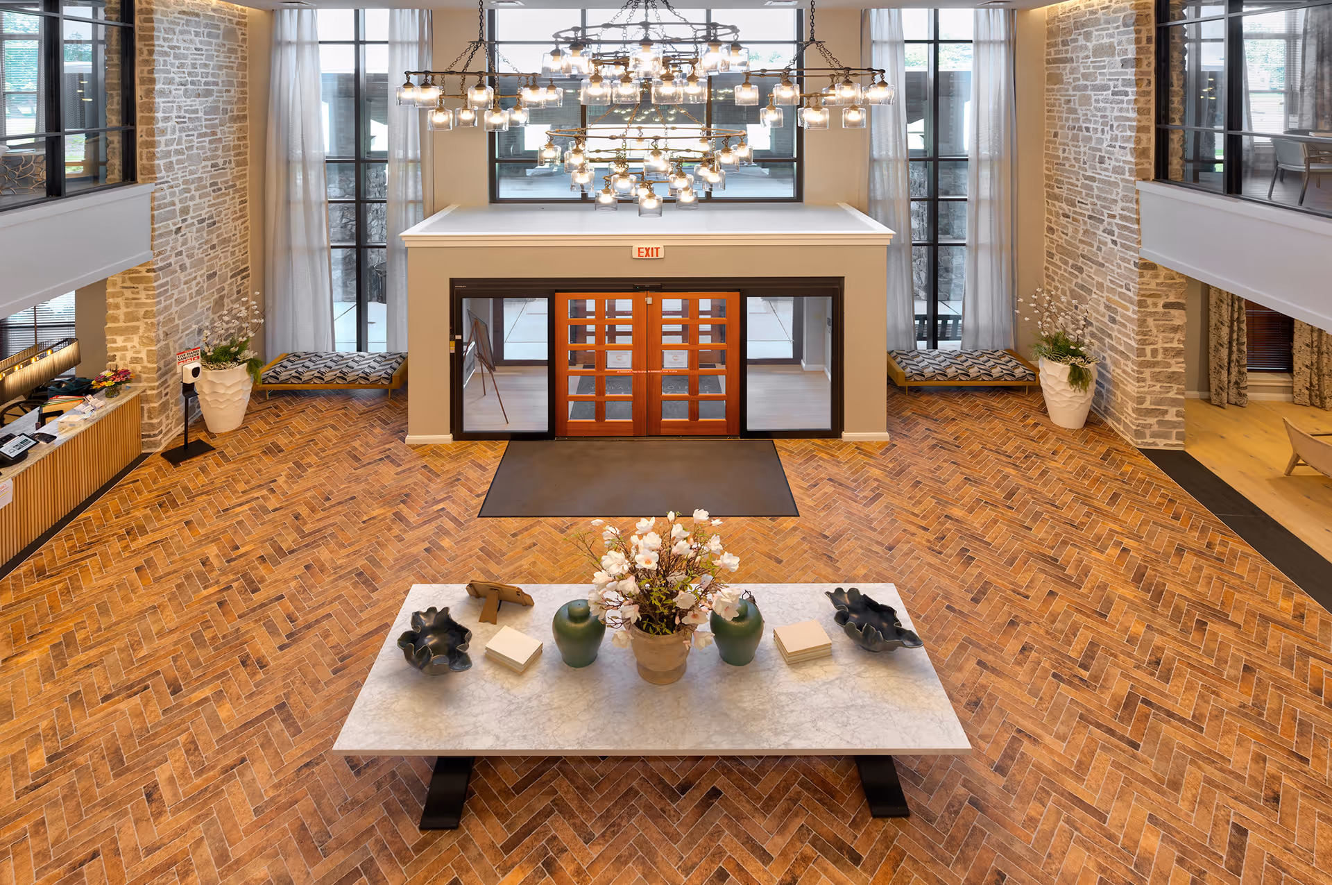 Spacious and elegant lobby area with a large chandelier hanging from the ceiling, a marble table with decorative vases and flowers in the center, wooden herringbone patterned floor, stone accent walls, large windows with sheer curtains, and double wooden doors marked with an exit sign.
