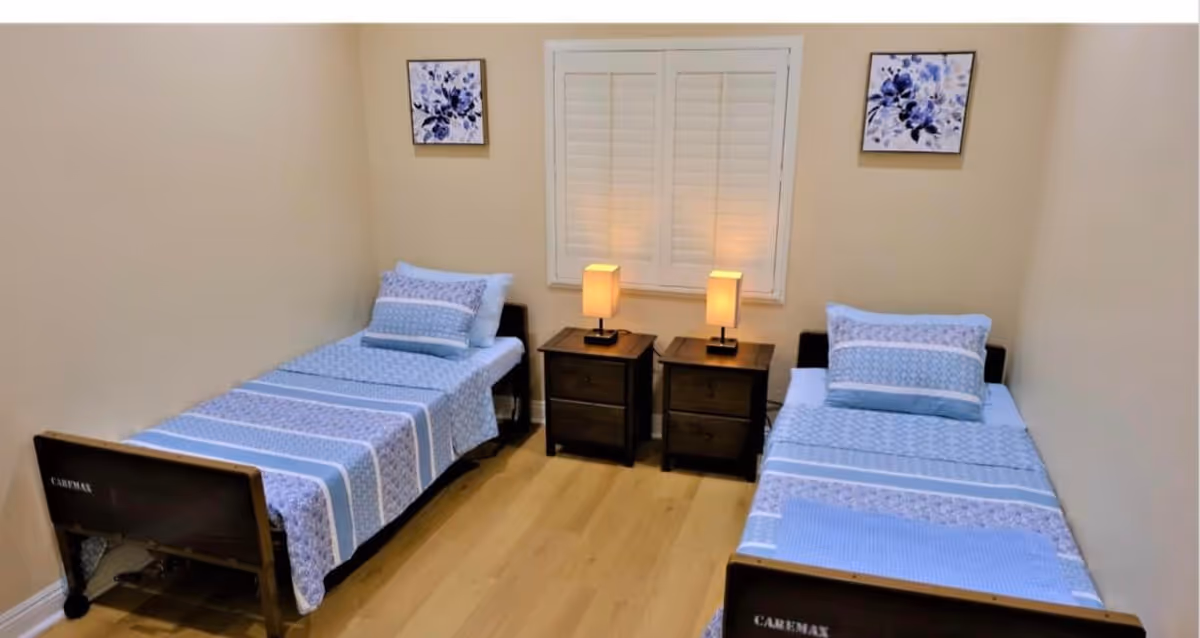 A simple bedroom with two single beds placed parallel to each other, each with blue patterned bedding and pillows. Between the beds are two dark wooden nightstands with matching lamps. The wall behind the beds has a window with closed white shutters and two framed floral artworks on either side.