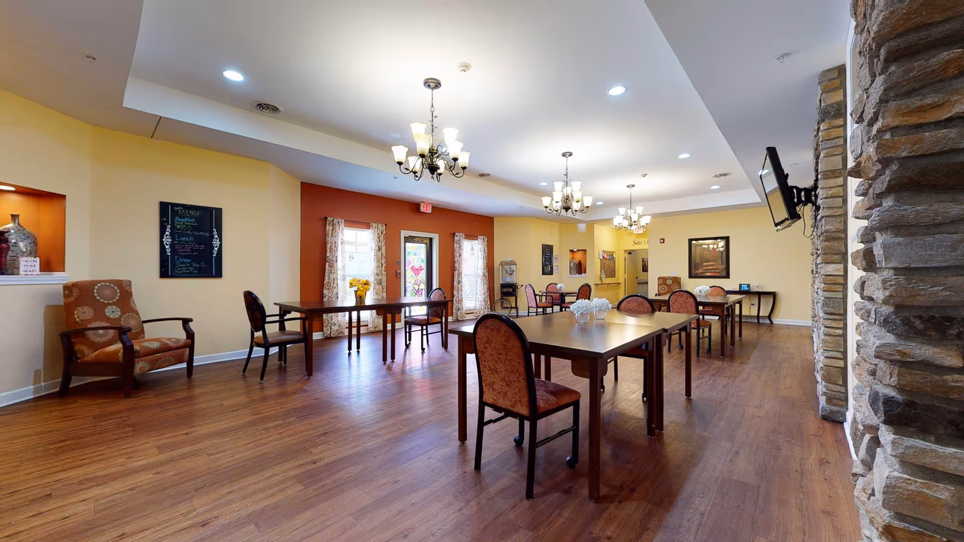 A spacious dining room with wooden floors, multiple tables and chairs arranged for seating, chandeliers hanging from the ceiling, a stone accent wall with a mounted TV, and windows with floral curtains allowing natural light to enter.