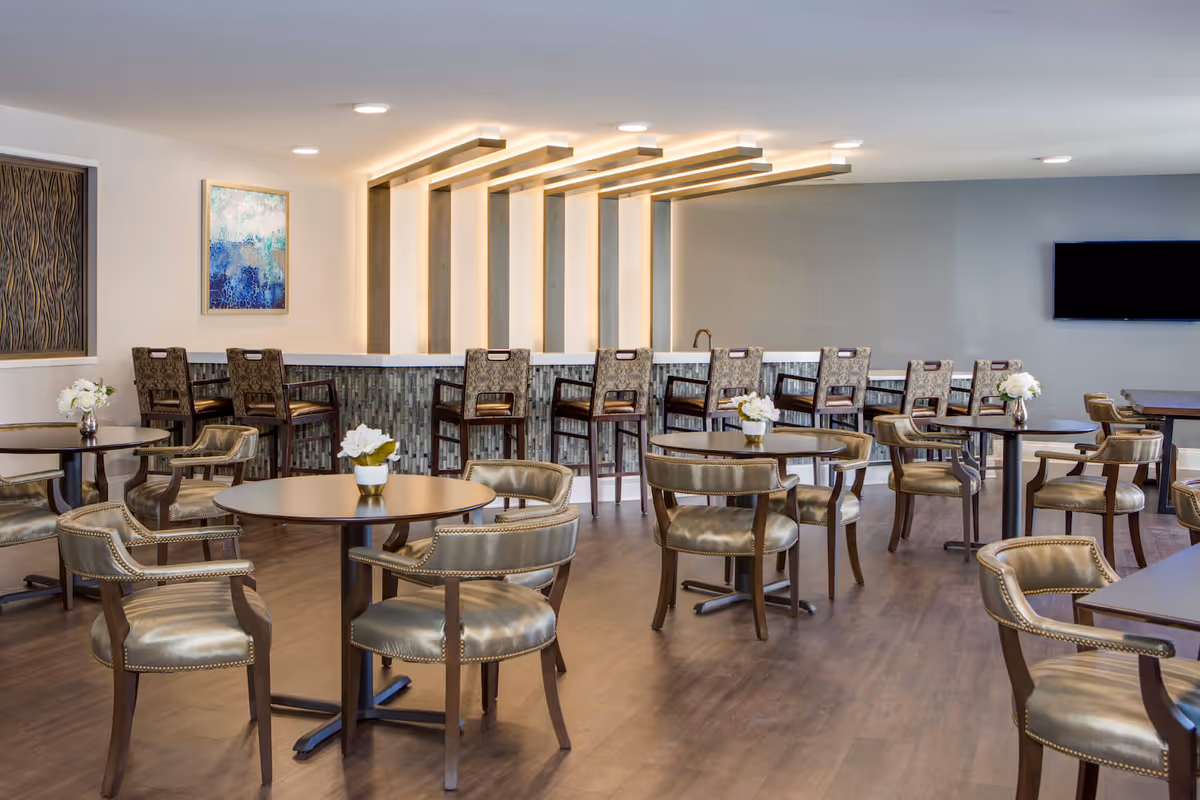 A modern dining area in a senior living facility with round tables and cushioned chairs arranged neatly. There is a bar counter with high chairs along the back wall, featuring vertical light fixtures above it. The room has wooden flooring, a wall-mounted TV, and decorative artwork on the walls.