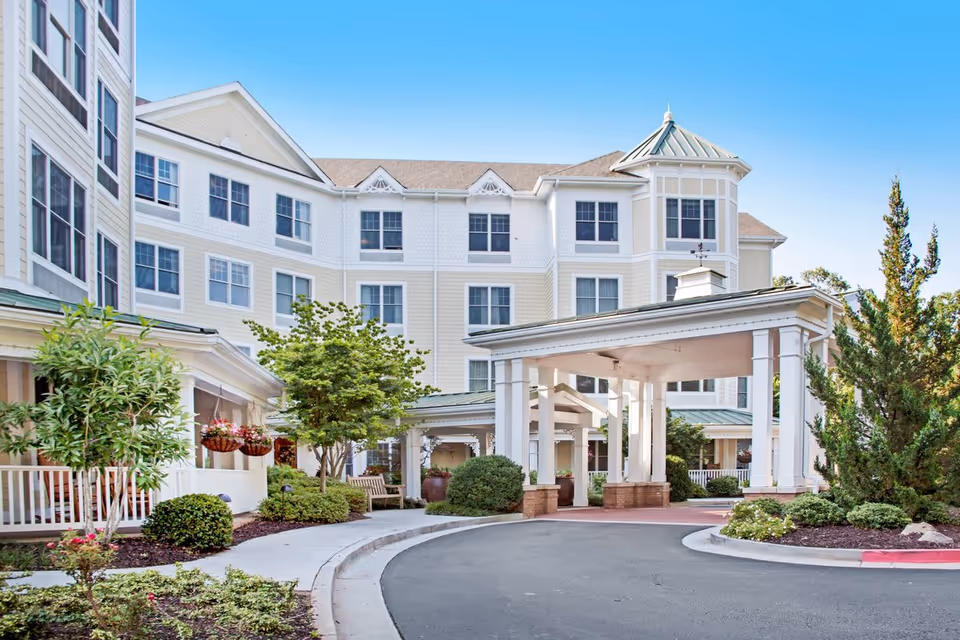 Front entrance and façade of a multi-story senior living building with a covered porte-cochere, porch, and landscaped driveway.