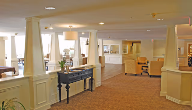Spacious interior common area with beige carpeting, white pillars, a black console table with decorative items, and seating areas with armchairs. Large windows allow natural light to fill the space.