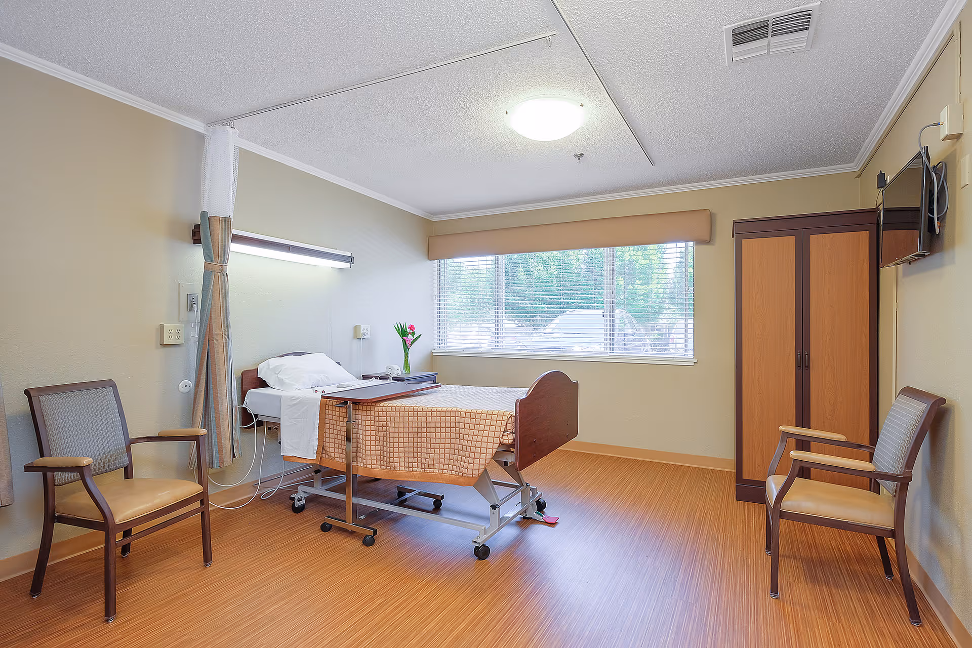 A clean and well-lit rehabilitation room with a hospital bed covered in a checkered blanket, two chairs on either side, a wooden wardrobe, a wall-mounted TV, and a window with blinds letting in natural light. A small vase with flowers is placed on the bedside table.