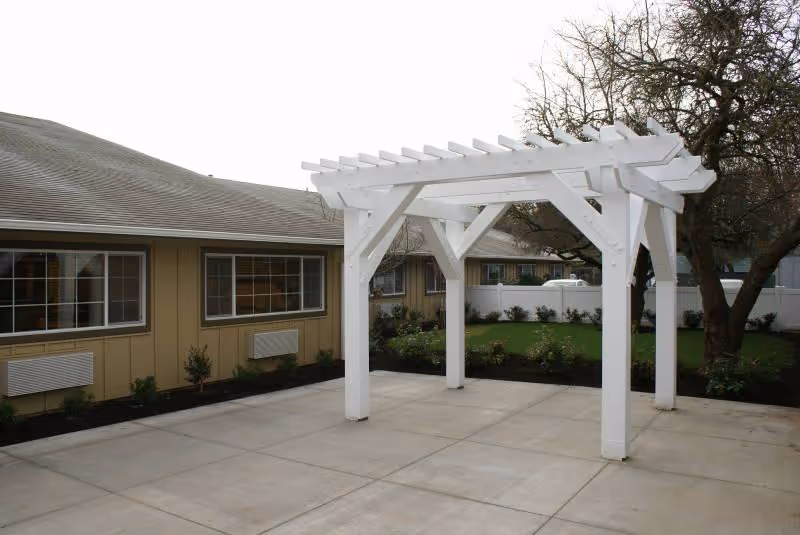 Outdoor patio area with a white wooden pergola structure on a concrete surface, surrounded by a garden with shrubs and trees. The background shows a beige building with multiple windows and a white fence enclosing the garden.