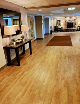 A spacious hallway with wooden flooring leading to a cozy seating area with two chairs and a stone fireplace. On the left side of the hallway, there is a black console table with two lamps, decorative plants, and a large mirror above it. The walls are painted in neutral tones, and the ceiling has recessed lighting.