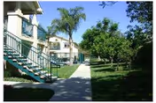 Outdoor view of a senior living facility with a paved walkway, green grass, palm trees, and other trees. There are two-story buildings with balconies and staircases on the left side of the walkway.
