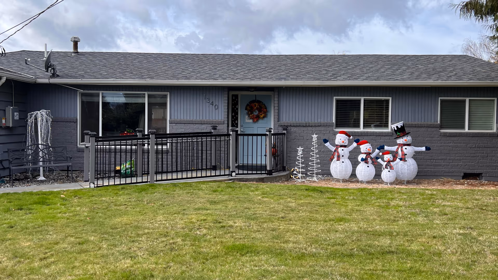 Front exterior view of a single-story gray house with a light blue door decorated with a wreath. There is a black metal railing along a small ramp leading to the door. Four inflatable snowmen and two white spiral Christmas tree decorations are displayed on the right side of the entrance. The lawn in front is green and well-kept, and the sky is cloudy.