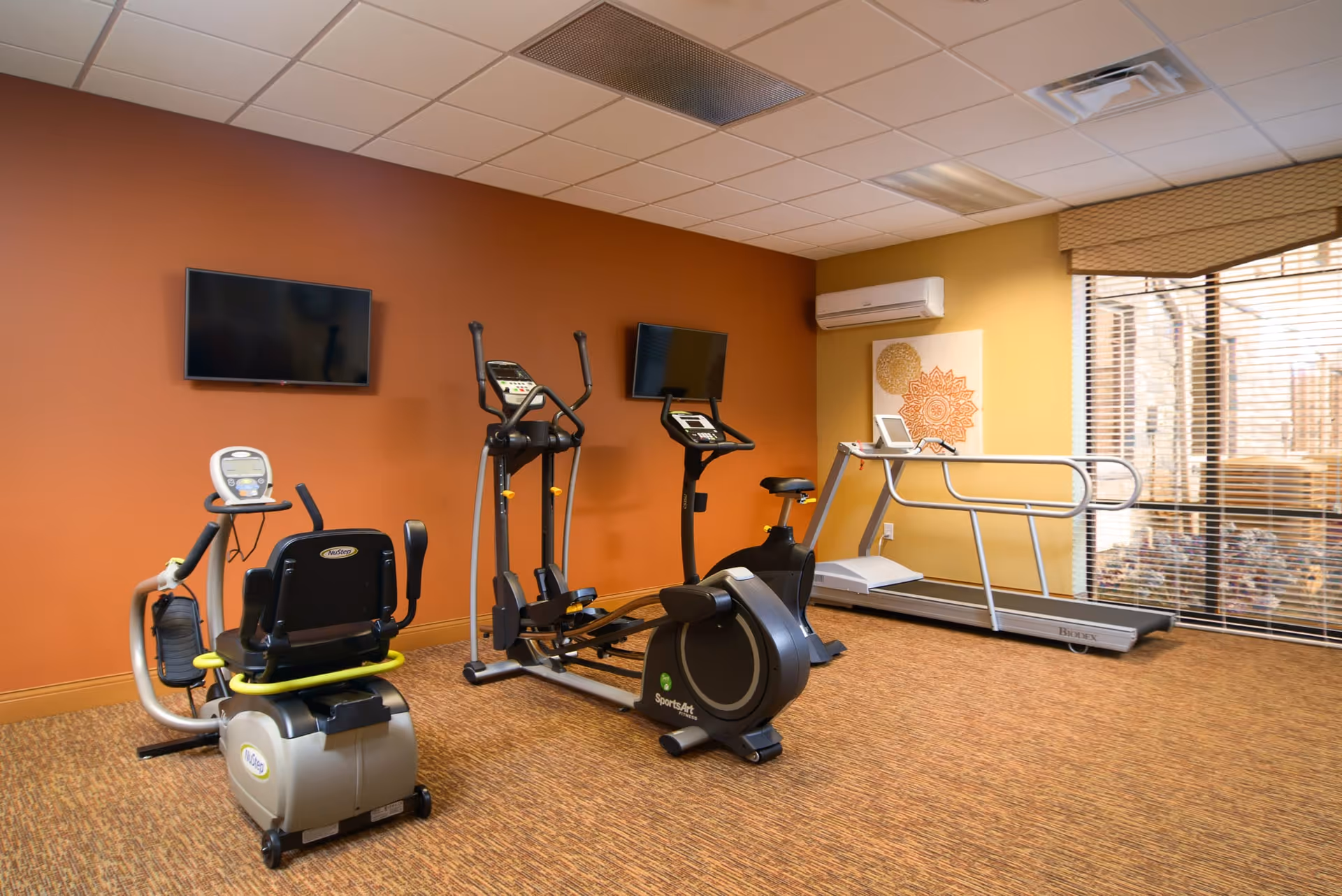 A small fitness room with exercise equipment including a recumbent bike, elliptical machine, stationary bike, and treadmill. The room has an orange accent wall with two mounted flat-screen TVs, a beige carpet, and a window with blinds letting in natural light. There is also a wall-mounted air conditioning unit and decorative artwork on the wall.