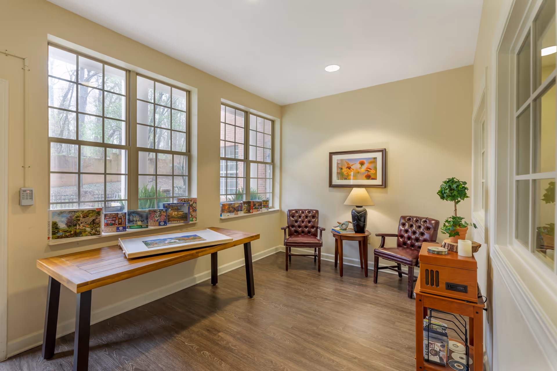 A bright room with large windows showing an outdoor view. Inside, there is a wooden table with several puzzle boxes on the windowsill, two brown leather tufted chairs with a small round table between them holding a lamp and some books. A framed picture of flowers hangs on the wall above the chairs. There is also a small wooden cabinet with a potted plant and some CDs on the right side.
