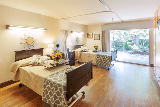 A bright and spacious room with two hospital-style beds, each with beige bedding and decorative pillows. Each bed has a bedside table with a lamp and a vase of flowers. The room features wooden flooring, wall-mounted lights, and a large sliding glass door that opens to a garden area outside. The walls are decorated with framed pictures and decorative wall hangings.