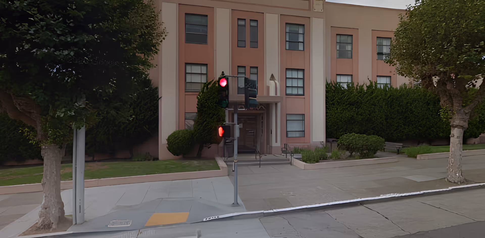 Exterior view of a multi-story building with pink and beige walls, several windows, and an entrance with a small ramp and stairs. There are green bushes and trees on either side of the entrance, and a traffic light showing red is visible in front of the building.