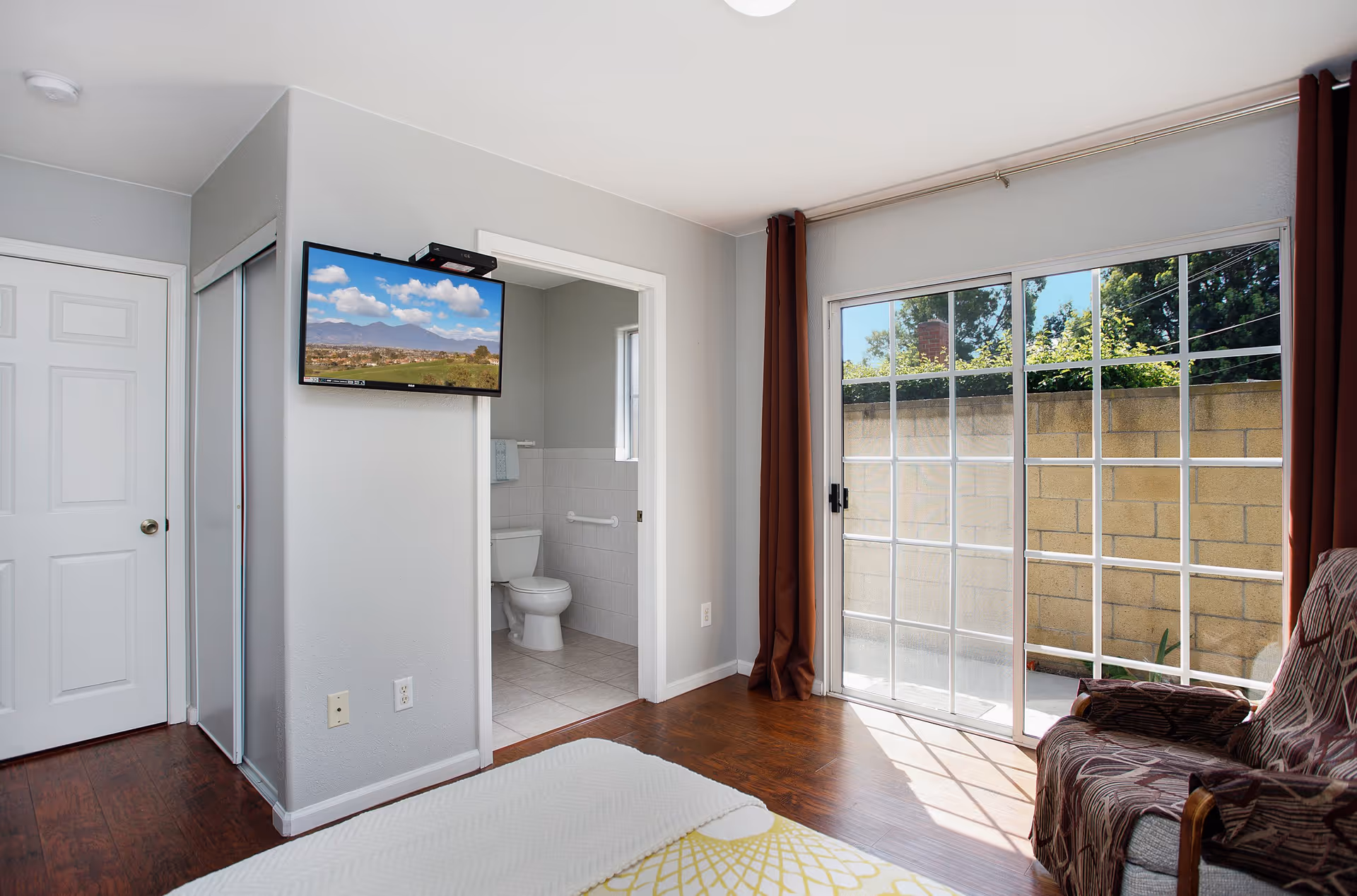A bright bedroom with wooden floors, a bed with a white and yellow patterned blanket, a wall-mounted TV showing a landscape, an armchair with a patterned cover, and a sliding glass door leading to an outdoor area. A bathroom with a toilet and tiled walls is visible through an open door.