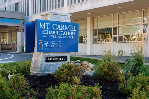 Blue sign reading "Mt. Carmel Rehabilitation and Nursing Center" and address in front of the facility building with landscaping in the foreground.