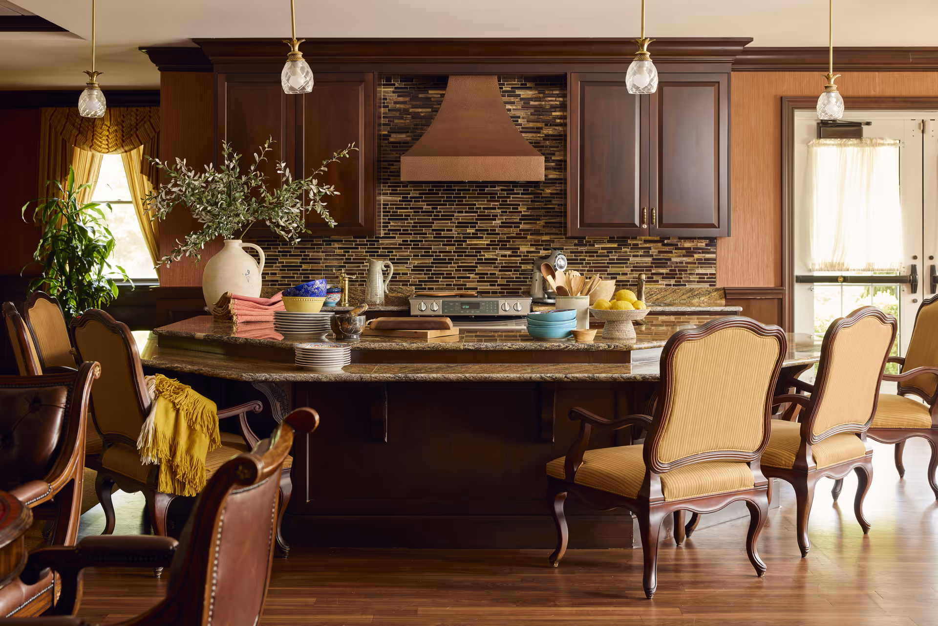 A warm, inviting kitchen area with a large granite island countertop surrounded by elegant wooden chairs with yellow upholstery. The kitchen features dark wood cabinets, a mosaic tile backsplash, a copper range hood, and various kitchenware including bowls, plates, and utensils on the counter. There are pendant lights hanging from the ceiling and a window with yellow curtains letting in natural light.